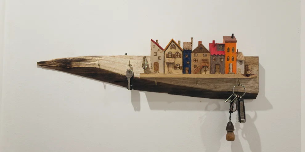 Porta-chaves de parede: uma tábua de madeira reciclada com bordo natural, encimada por casinhas em miniatura esculpidas e pintadas à mão e cinco ganchos metálicos