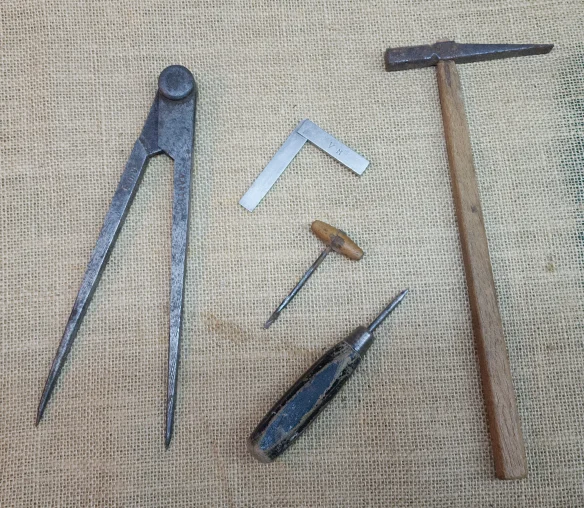 Quatro ferramentas manuais antigas sobre juta: um compasso, um esquadro, uma sovela e um martelo de pena cruzada